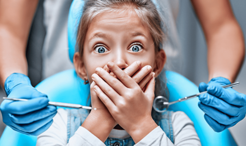 Anxious child covering mouth with hands at dental appointment with dentist holding instruments.