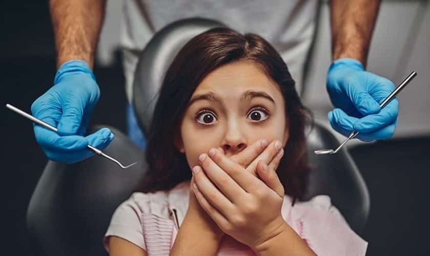 Young girl with anxious expression covering mouth while dentist holds dental tools in operatory.